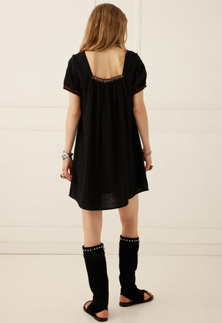 a-woman-viewed-from-behind-wearing-a-loose-black-short-sleeve-dress-with-a-decorative-embroidered-neckline-black-studded-leg-warmers-and-black-sandals-standing-against-a-white-paneled-wall
