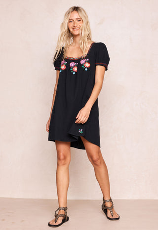 a-woman-modeling-a-black-short-sleeve-mini-dress-with-colorful-floral-embroidery-around-the-neckline-paired-with-brown-strappy-sandals-against-a-textured-beige-studio-backdrop