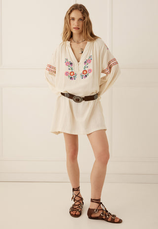 a-woman-modeling-a-cream-colored-bohemian-mini-dress-with-colorful-floral-embroidery-on-the-chest-and-sleeves-styled-with-a-brown-belt-and-brown-lace-up-sandals-against-a-cream-paneled-wall