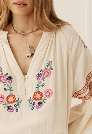 a-close-up-of-a-woman-wearing-a-cream-colored-bohemian-blouse-with-colorful-floral-embroidery-on-the-front-and-sleeves-accessorized-with-layered-silver-necklaces