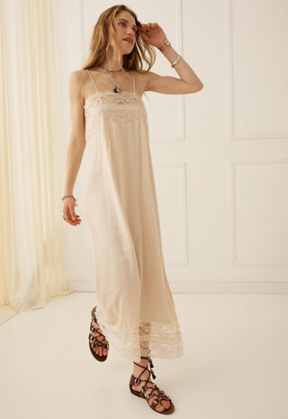 a-woman-models-a-long-cream-colored-slip-dress-with-delicate-lace-trim-at-the-bodice-and-hem-paired-with-brown-strappy-flat-sandals-in-a-bright-minimalist-room-with-white-paneled-walls-and-a-sheer-cream-curtain