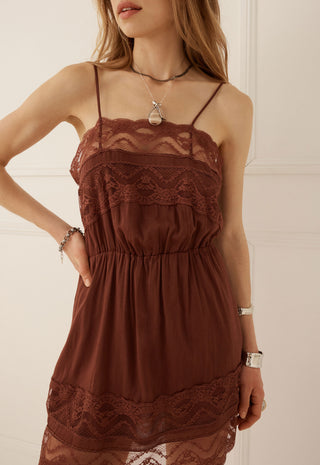 a-woman-wearing-a-brown-lace-trimmed-spaghetti-strap-camisole-dress-with-a-gathered-empire-waist-shown-from-the-neck-down-against-a-light-cream-paneled-wall