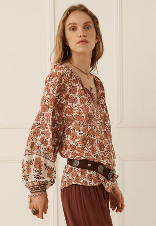 a-woman-wearing-a-bohemian-style-long-sleeve-blouse-with-a-rust-and-beige-floral-print-paired-with-a-dark-brown-belt-and-matching-brown-skirt-standing-against-a-white-paneled-wall