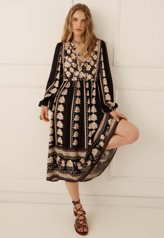 a-woman-modeling-a-black-midi-dress-with-beige-floral-embroidery-and-lace-detailing-paired-with-brown-strappy-lace-up-sandals-standing-against-a-white-paneled-wall
