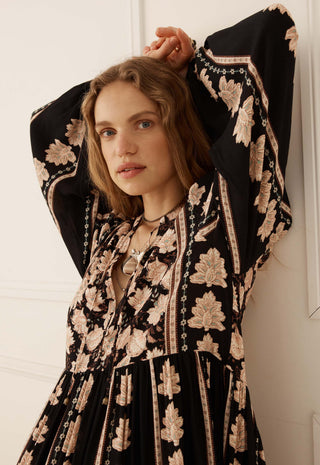 a-woman-wearing-a-black-bohemian-style-maxi-dress-with-beige-floral-embroidery-and-wide-bell-sleeves-posing-against-a-white-paneled-wall