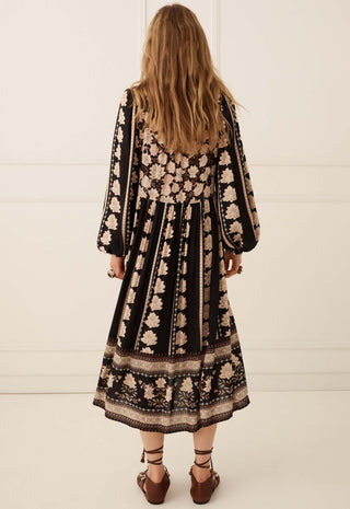 a-woman-viewed-from-behind-wearing-a-long-sleeved-midi-dress-with-a-black-base-and-beige-floral-pattern-paired-with-brown-lace-up-shoes-set-against-a-clean-white-paneled-wall