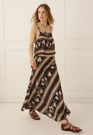 a-woman-modeling-a-two-piece-bohemian-maxi-set-with-a-black-brown-and-beige-striped-pattern-featuring-a-spaghetti-strap-crop-top-and-a-long-skirt-paired-with-brown-strappy-sandals-against-a-white-paneled-wall