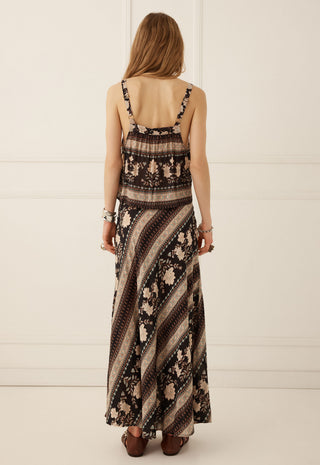 a-woman-viewed-from-behind-wearing-a-sleeveless-maxi-dress-with-a-brown-beige-and-black-striped-and-floral-pattern-paired-with-brown-flat-shoes-against-a-white-paneled-wall