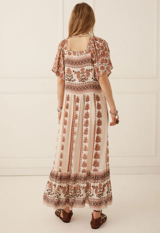 a-woman-viewed-from-behind-wearing-a-bohemian-style-maxi-dress-with-brown-and-beige-patterns-standing-against-a-white-paneled-wall