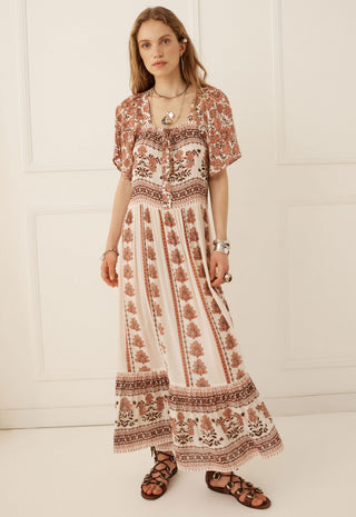 a-woman-modeling-a-bohemian-style-maxi-dress-with-a-brown-and-beige-floral-pattern-standing-against-a-white-paneled-wall