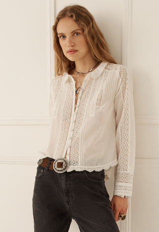a-woman-wearing-a-white-long-sleeve-blouse-with-intricate-lace-and-embroidered-detailing-tucked-into-dark-gray-jeans-with-a-brown-belt-standing-against-a-white-paneled-wall