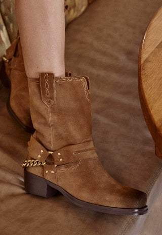 a-close-up-of-a-persons-leg-wearing-a-brown-suede-western-style-boot-with-a-dark-brown-block-heel-decorative-stitching-and-a-gold-chain-detail-on-the-side-set-against-a-brown-leather-couch