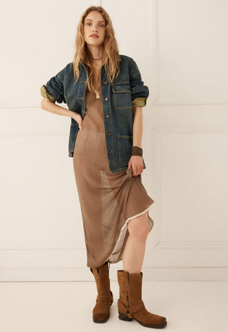 a-woman-models-a-bohemian-style-outfit-featuring-a-brown-silk-slip-dress-a-blue-denim-jacket-with-olive-green-rolled-up-sleeves-and-brown-suede-boots-set-against-a-clean-white-paneled-wall