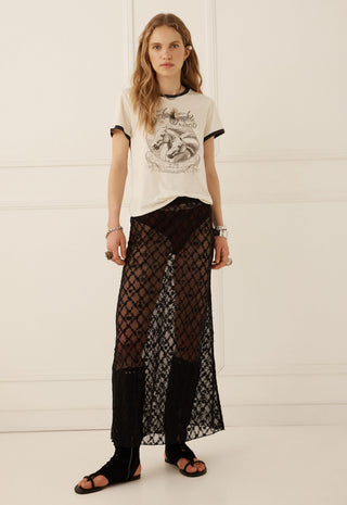 a-model-stands-against-a-white-paneled-wall-wearing-a-cream-graphic-t-shirt-with-black-trim-sheer-black-patterned-maxi-pants-and-black-sandals