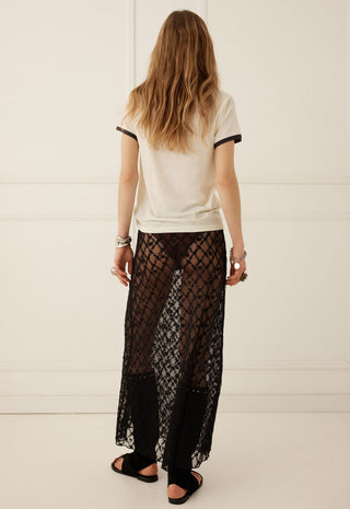 a-woman-viewed-from-behind-wearing-a-cream-ringer-t-shirt-with-black-trim-and-a-sheer-black-lace-maxi-skirt-standing-against-a-white-paneled-wall