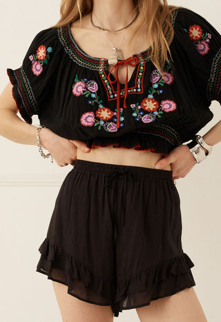 a-woman-wearing-a-black-embroidered-crop-top-with-colorful-floral-patterns-and-matching-black-ruffled-shorts-standing-against-a-light-beige-wall